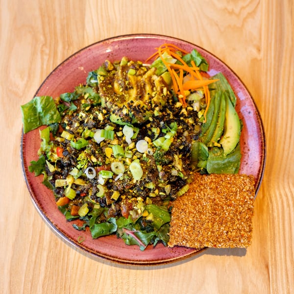 Colorful vegan salad with avocado, pistachios, black sesame seeds, and a seed cracker on a pink plate.