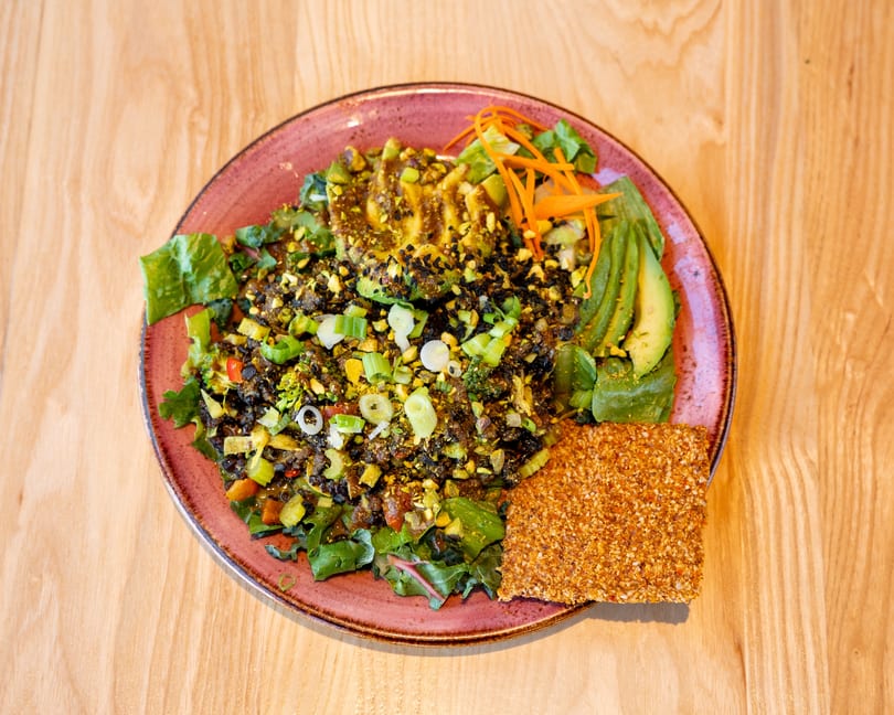 Colorful vegan salad with avocado, pistachios, black sesame seeds, and a seed cracker on a pink plate.