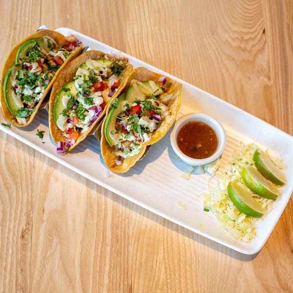 Three tacos with avocado, salsa, and cilantro, served with lime wedges and sauce on a white plate.