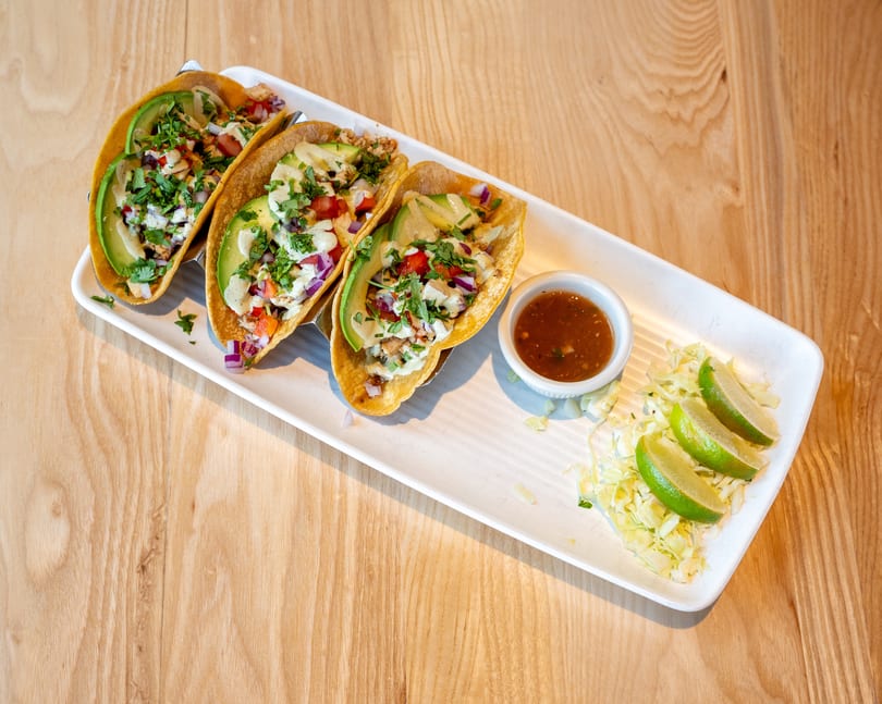 Three tacos with avocado, salsa, and cilantro, served with lime wedges and sauce on a white plate.