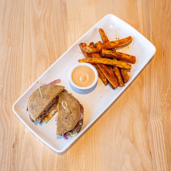 A burger cut in half with sweet potato fries and dipping sauce on a white rectangular plate.