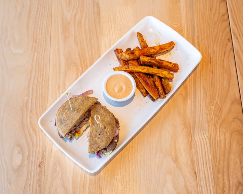 A burger cut in half with sweet potato fries and dipping sauce on a white rectangular plate.