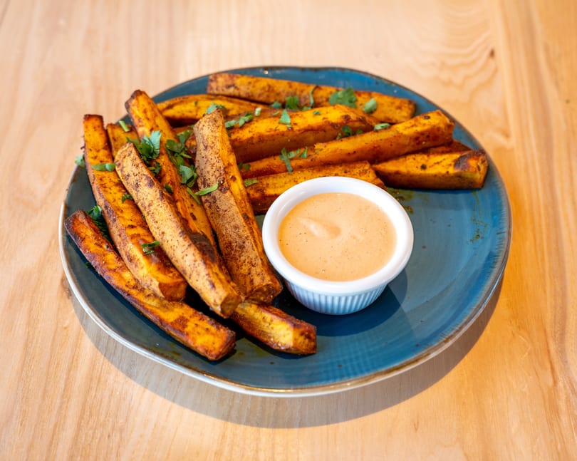 Sweet potato fries seasoned with herbs and spices, served with a side of dipping sauce on a blue plate.