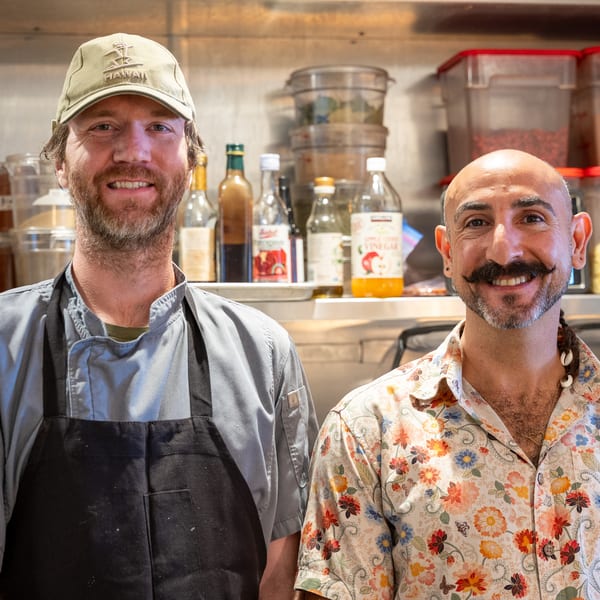 Two smiling men, one in a chef's coat and the other in a floral shirt, stand in a kitchen.