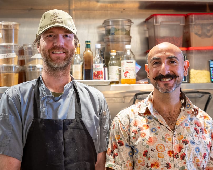 Two smiling men, one in a chef's coat and the other in a floral shirt, stand in a kitchen.