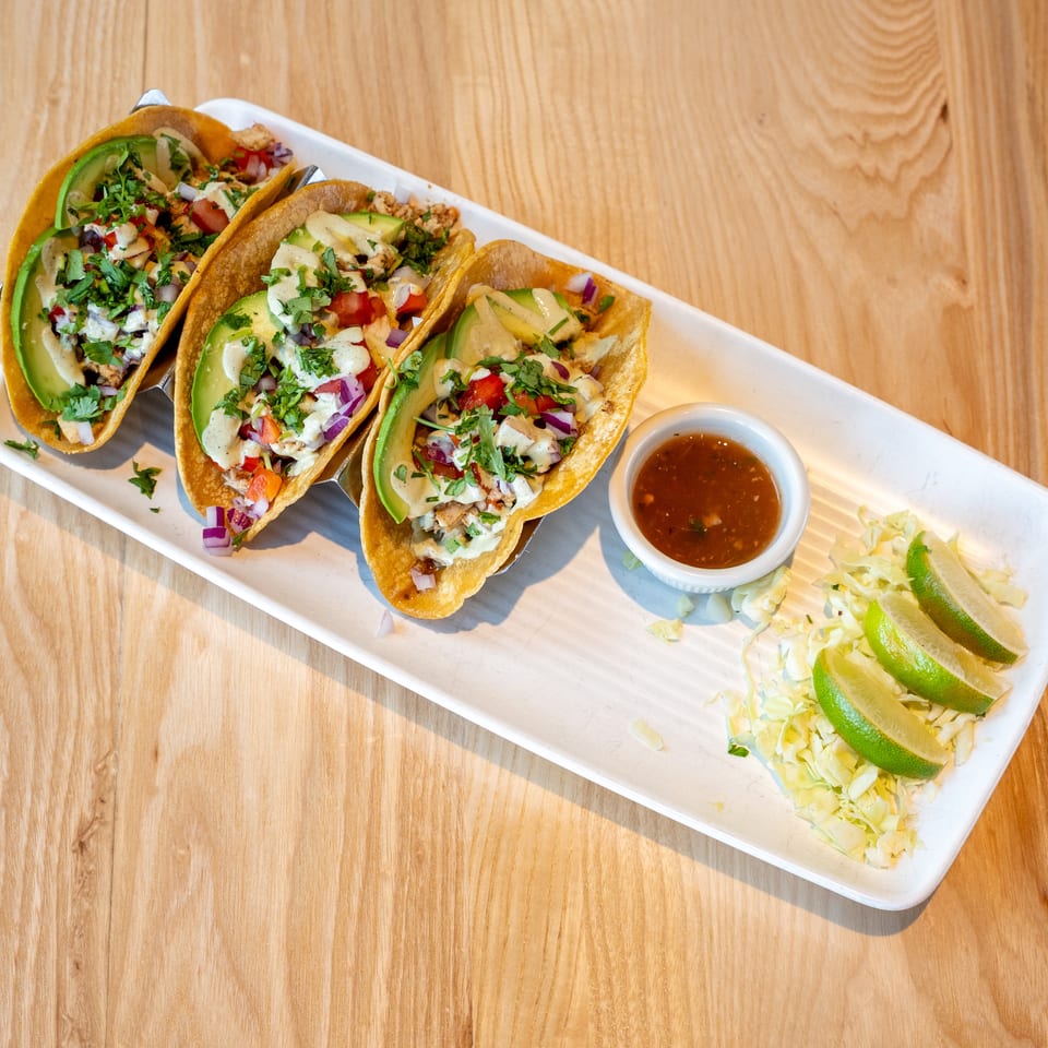 Three tacos with avocado, cilantro, and sauce on a white plate with salsa, shredded cabbage, and lime.
