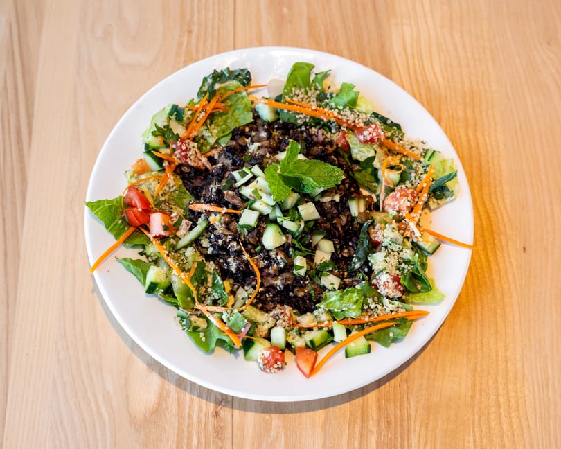 A vibrant salad with dark lentils, fresh greens, cucumbers, tomatoes, carrots, and mint on a white plate.