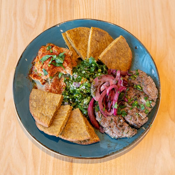 A blue plate with hummus, tabbouleh, grilled meat patties, pickled red onions, and pita bread.