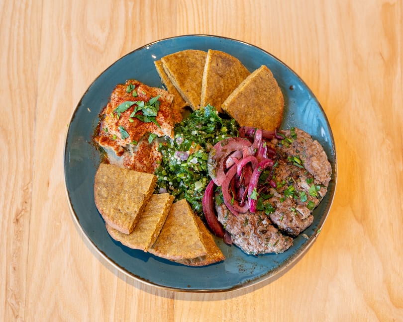 A blue plate with hummus, tabbouleh, grilled meat patties, pickled red onions, and pita bread.