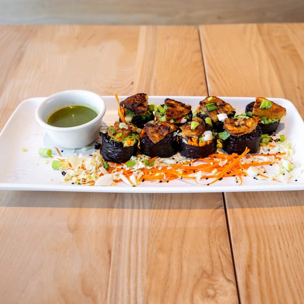 A white rectangular plate holds six dark sushi rolls topped with fried plantains and green onions, alongside a green dipping sauce.