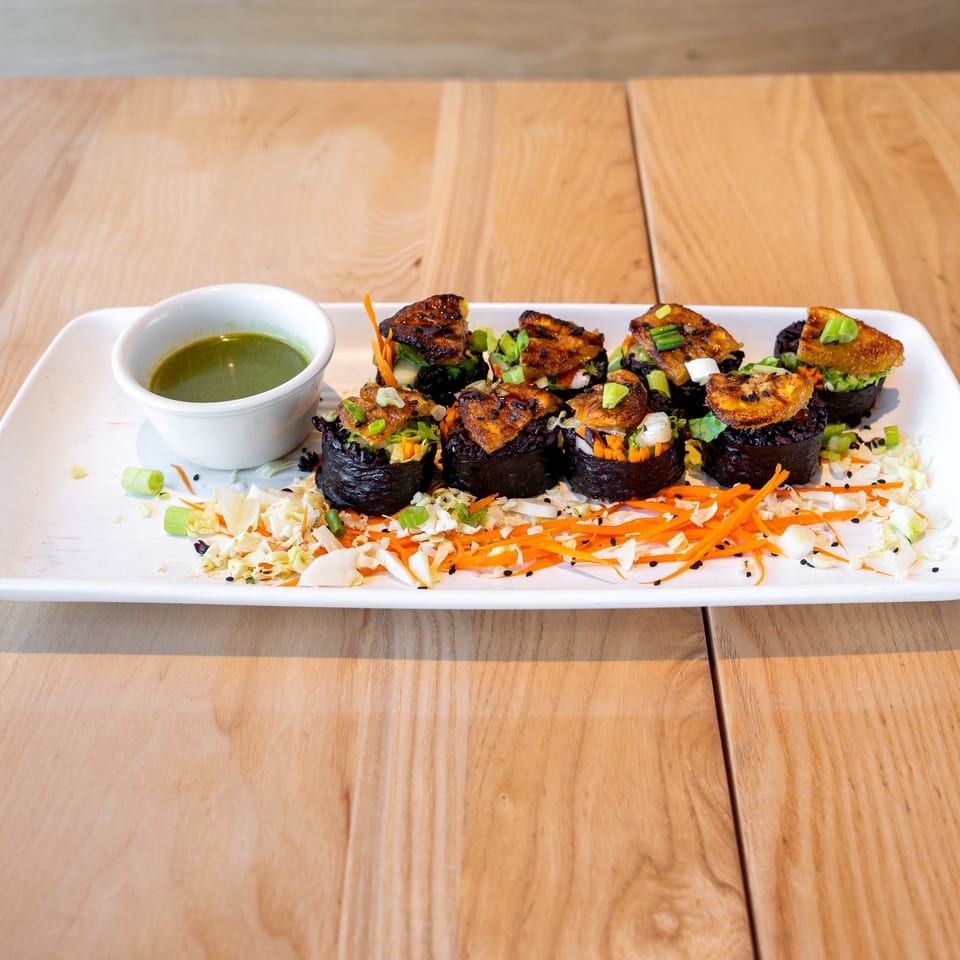 A white rectangular plate holds six dark sushi rolls topped with fried plantains and green onions, alongside a green dipping sauce.