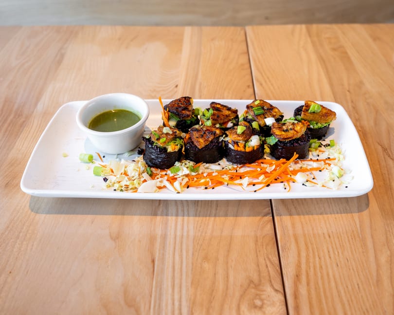 A white rectangular plate holds six dark sushi rolls topped with fried plantains and green onions, alongside a green dipping sauce.
