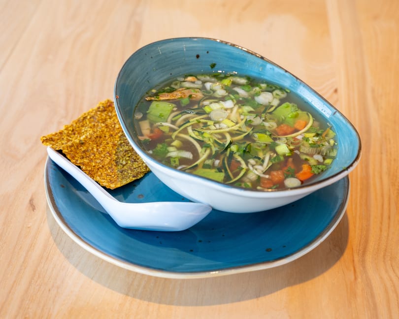 Clear vegetable soup with zucchini noodles, carrots, and scallions, served with a cracker.