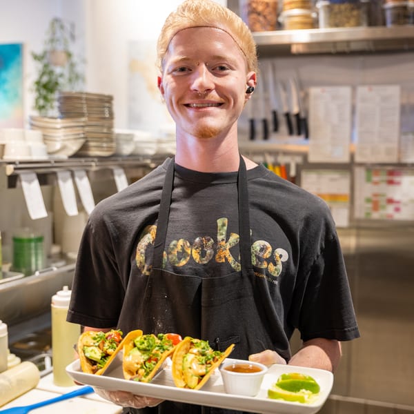 Smiling chef in a black apron holding a tray of three tacos with lime wedges and sauce.