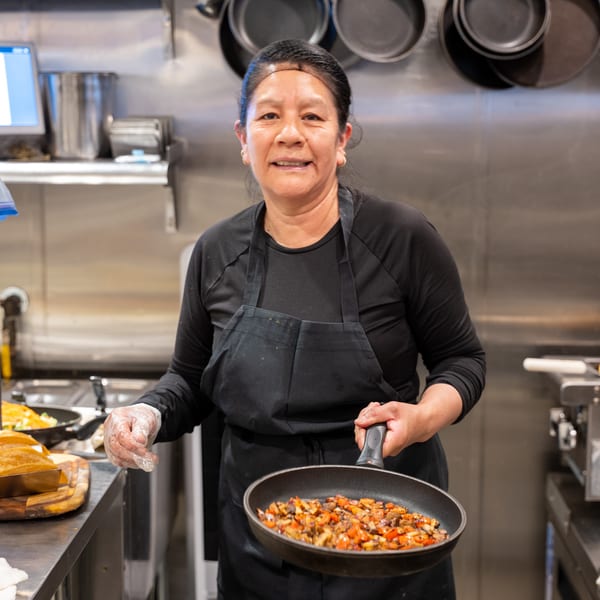 A smiling chef in a black apron holds a pan of sautéed vegetables in a commercial kitchen.