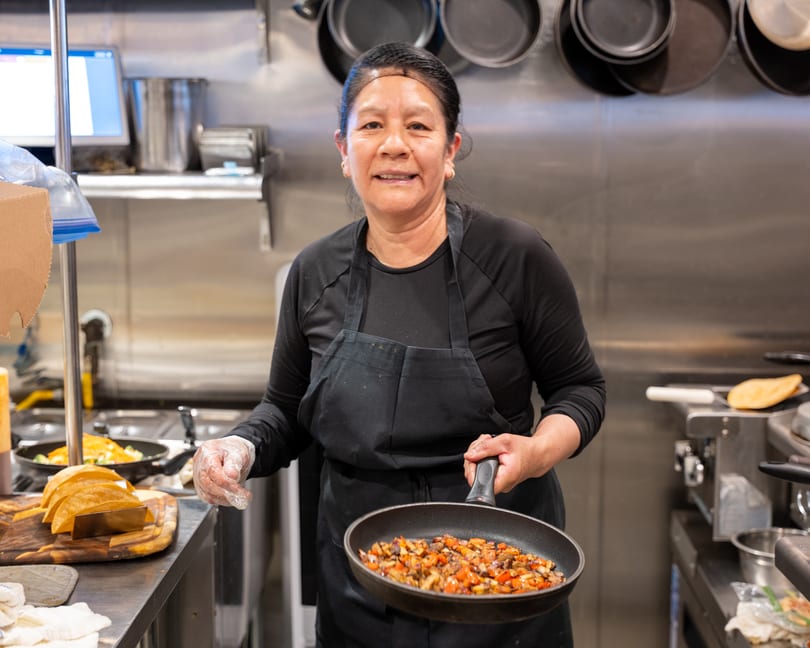 A smiling chef in a black apron holds a pan of sautéed vegetables in a commercial kitchen.