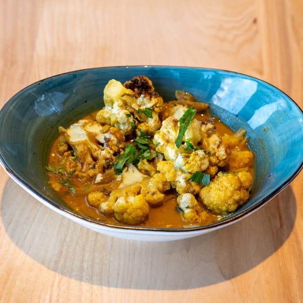 A blue bowl filled with cauliflower curry, garnished with fresh herbs, on a wooden table.