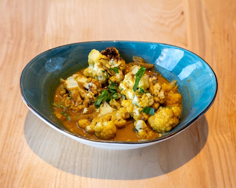 A blue bowl filled with cauliflower curry, garnished with fresh herbs, on a wooden table.