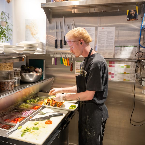 A chef in a commercial kitchen preparing tacos with fresh ingredients at a food prep station.