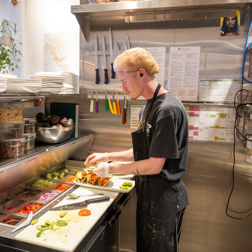A chef in a commercial kitchen preparing tacos with fresh ingredients at a food prep station.