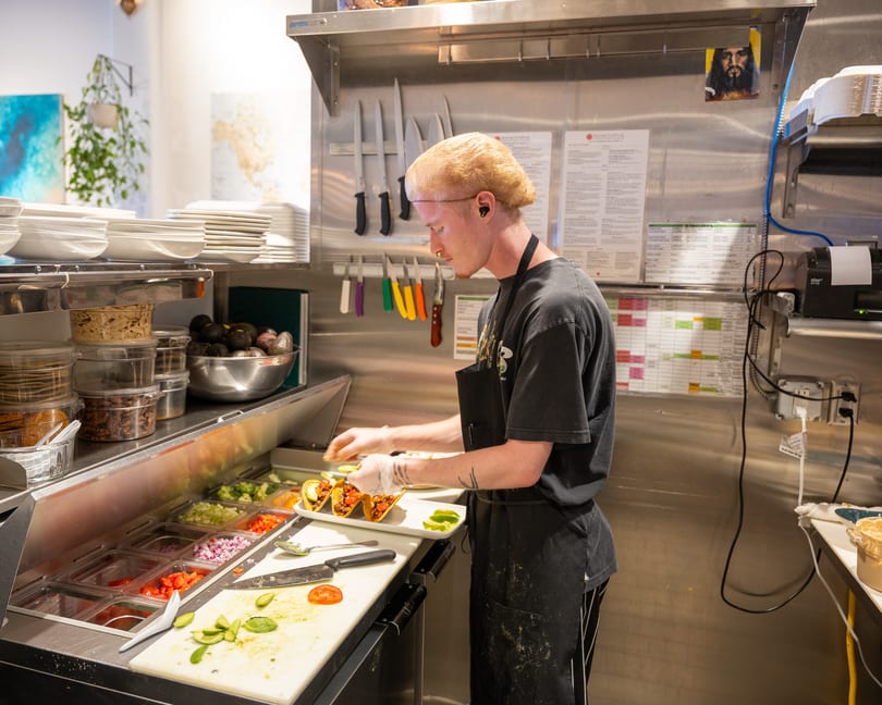 A chef in a commercial kitchen preparing tacos with fresh ingredients at a food prep station.