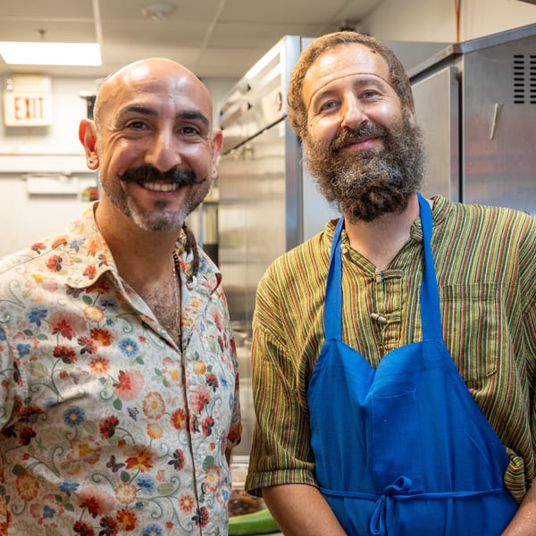 Two smiling chefs, one with a mustache and floral shirt, the other bearded in a striped shirt and blue apron.